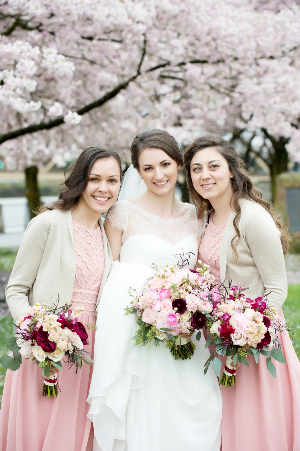 bridesmaids in pink dresses and tan cardigans hug around a bride holding pink and maroon bouquets with cherry blossom trees in the background