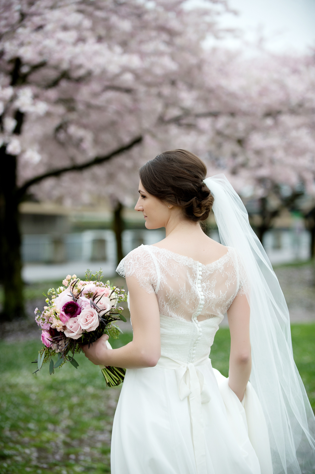a bride in a pretty lace wedding dress holds a pink and maroon bouquet in front of cherry blossom trees on the portland waterfront