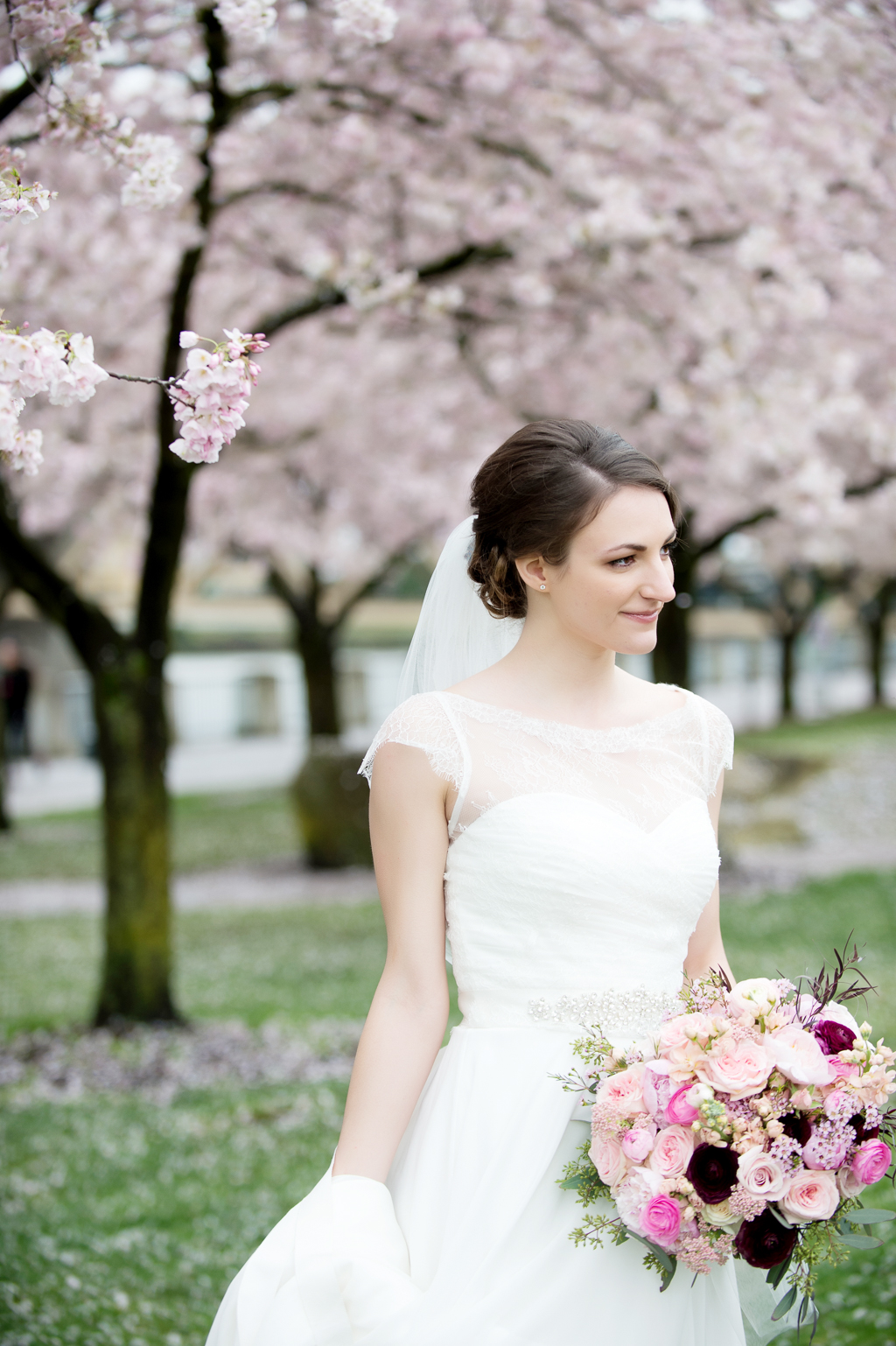 a bride in a pretty lace wedding dress holds a pink and maroon bouquet in front of cherry blossom trees on the portland waterfront
