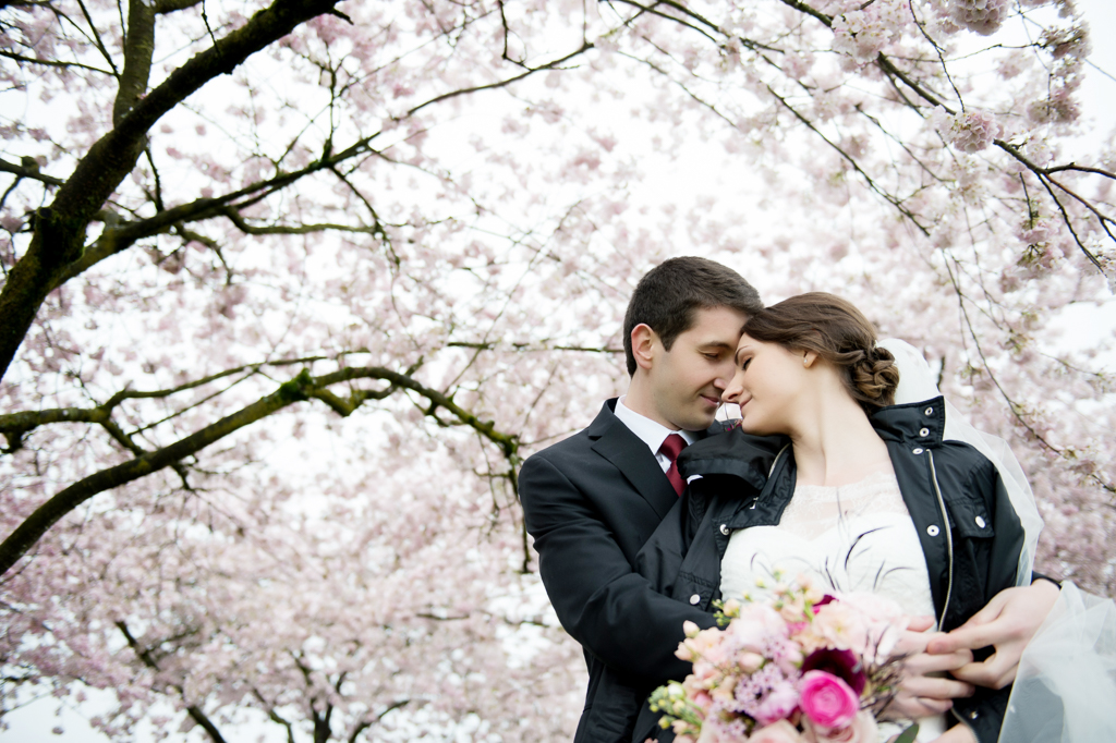 a bride wearing a black leather coat leans into her groom's arms underneath cherry blossom trees on the portland waterfront