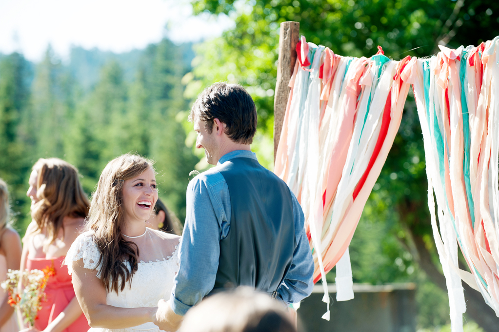 a bride and groom stand in front of a wedding arbor is hung with torn pieces of cloth in orange red peach and turquoise