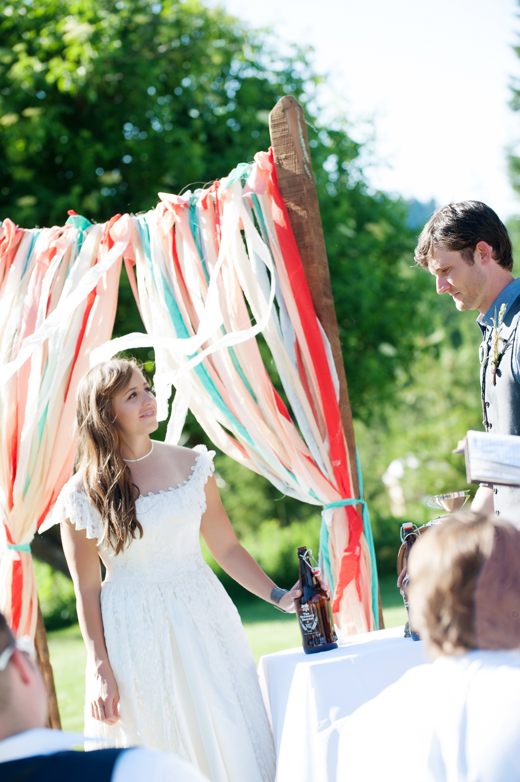 a bride and groom stand in front of a wedding arbor is hung with torn pieces of cloth in orange red peach and turquoise 