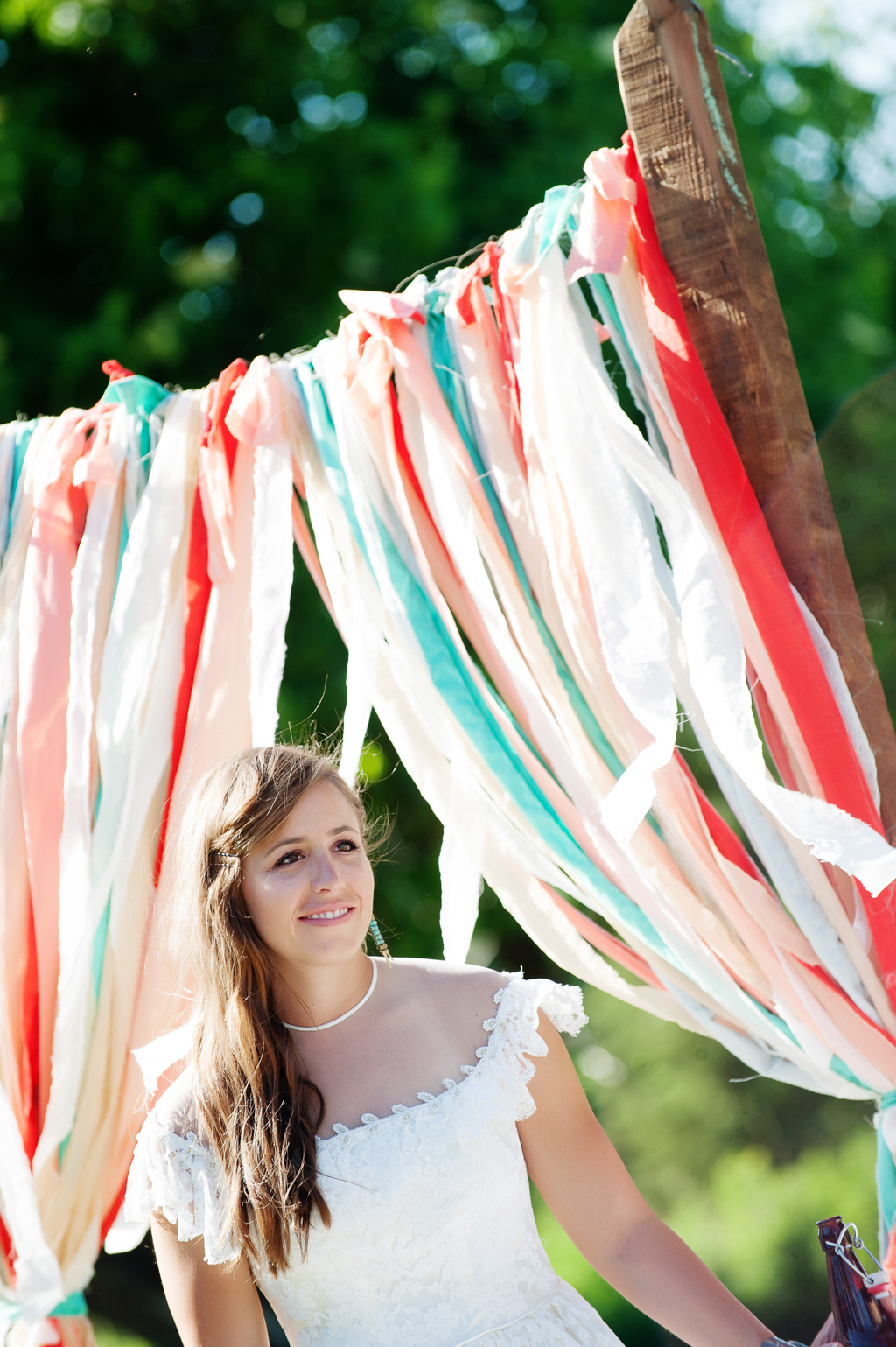 a bride stands in front of a wedding arbor is hung with torn pieces of cloth in orange red peach and turquoise 
