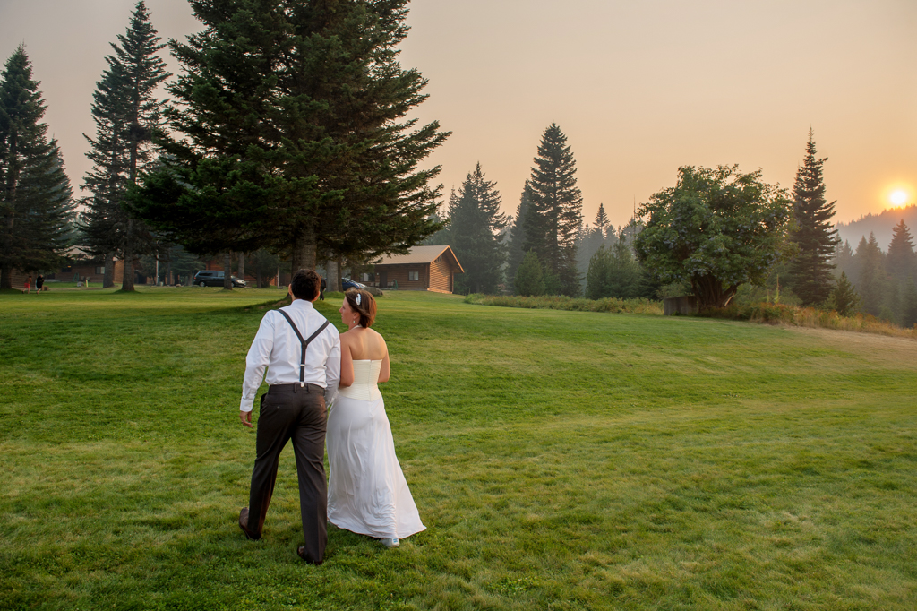 a bride and groom walks through the yard at mount hood at sunset with the sky filled with orange forest fire smoke and fog