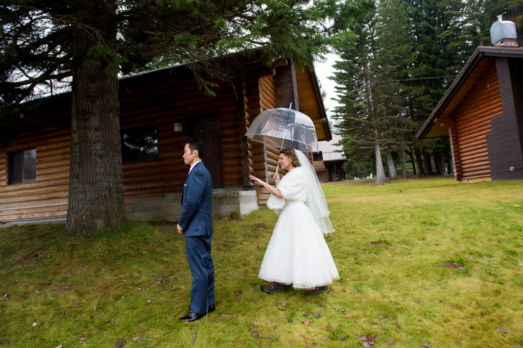 bride sneaks up behind groom holding a clear umbrella