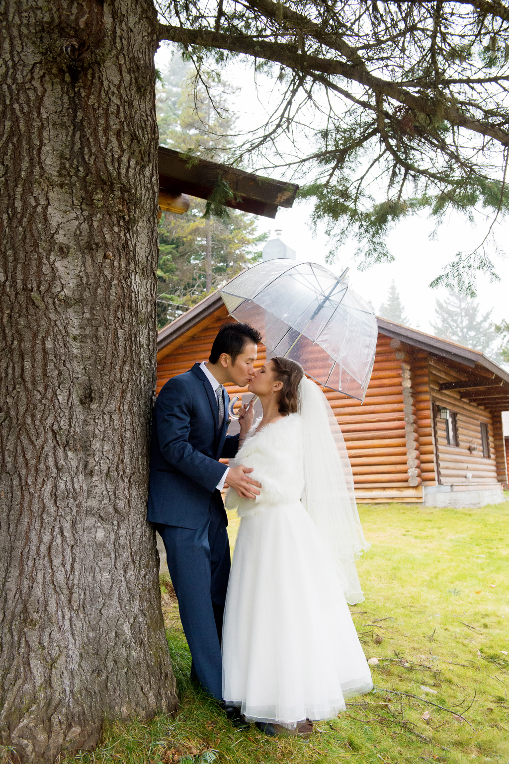groom leans against a tree holding a clear umbrella and the bride kisses him