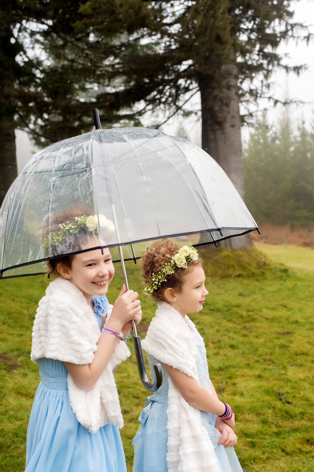 two flower girls wearing blue dresses and fur shawls stand under and umbrella
