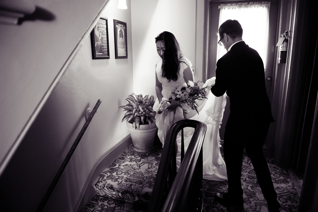 groom holds the bride's wedding dress as she walks down the stairs 