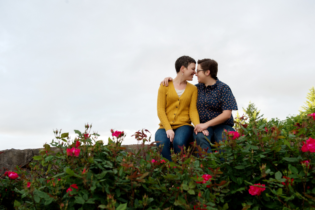 two girls sit on the wall and embrace in front of the roses at council crest park