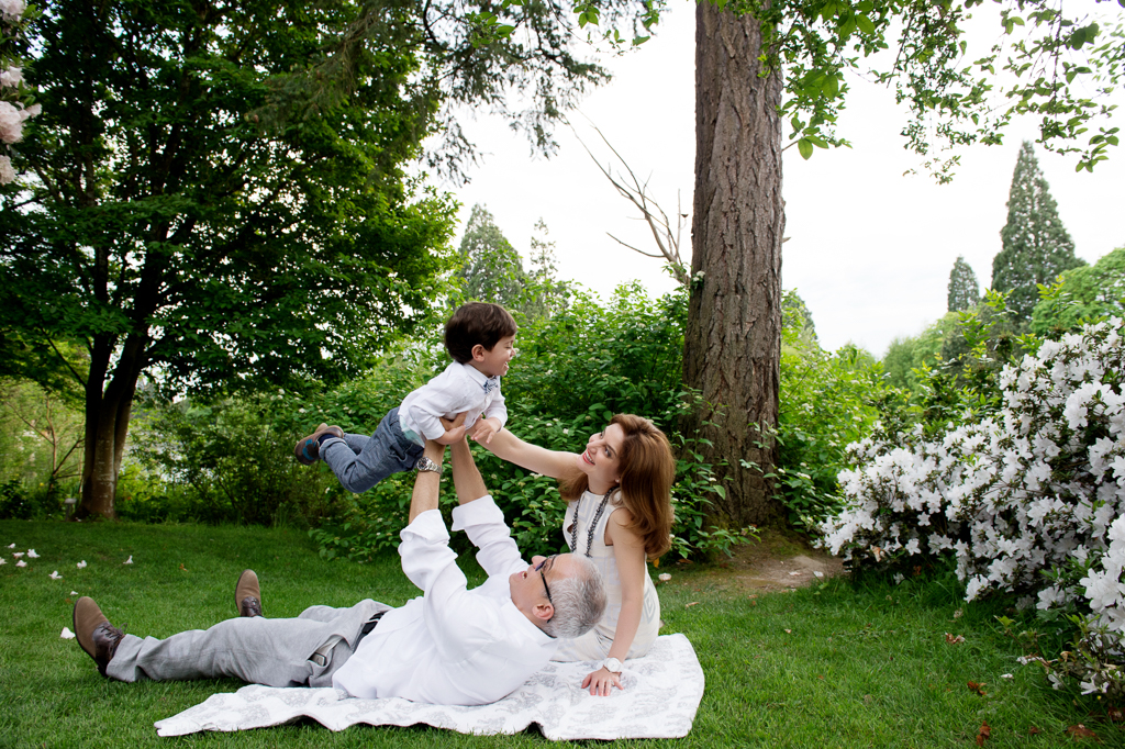 a dad lays in the grass and holds a little boy in a bowtie in the air while mom looks on