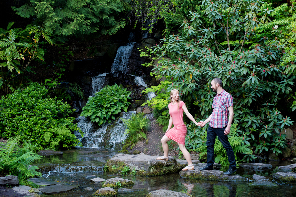 a girl pulls a boy by his hand across rocks in front of a waterfall