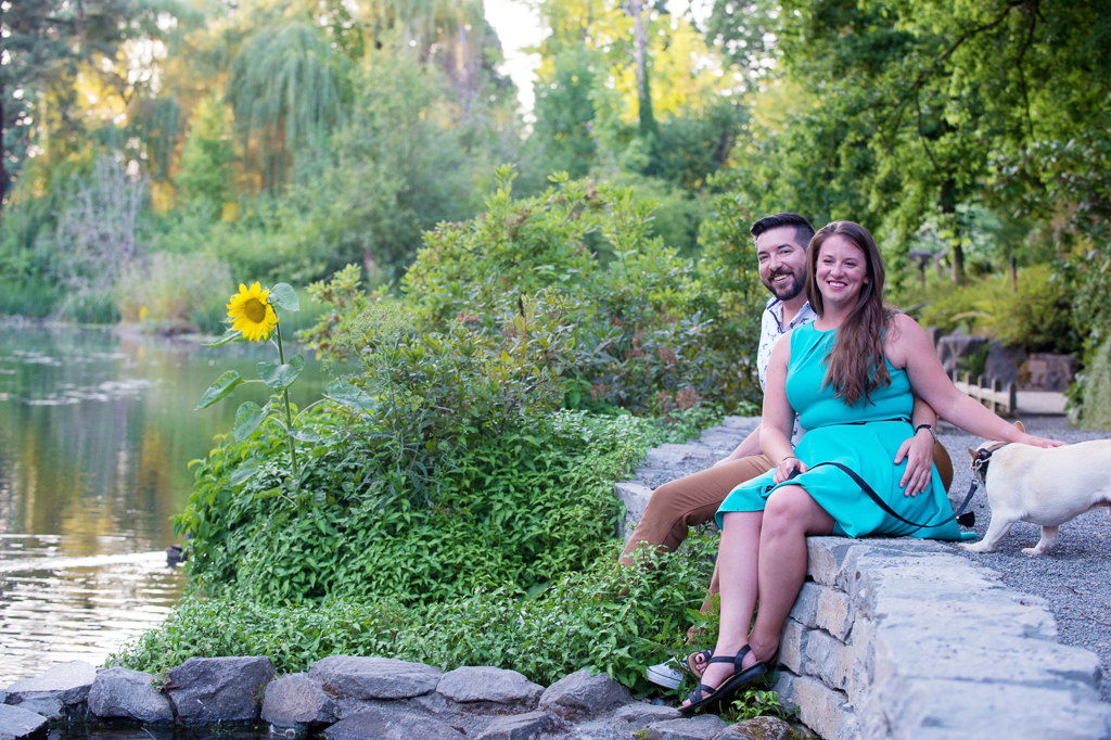 a couple sit on a stone wall with one sunflower growing behind them