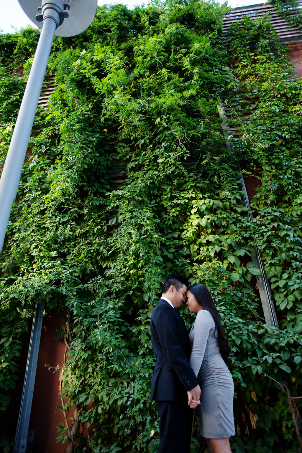 a couple stand with their heads together in front of a tall wall of ivy