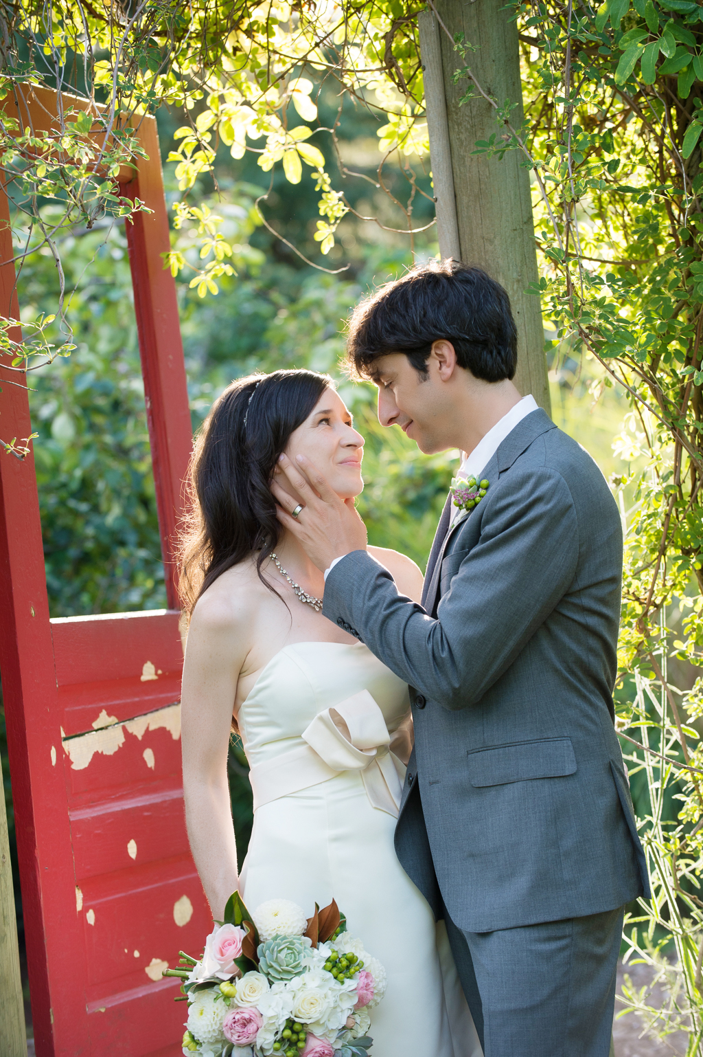 groom caresses bride's cheek in garden with red door at edgefield