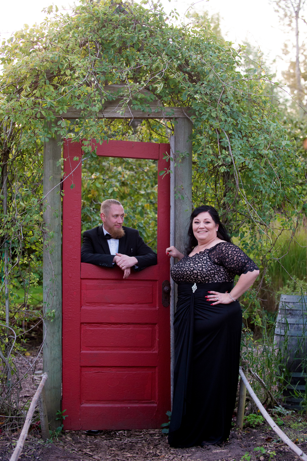 a bride wears a black wedding dress stands with her groom by a red door in a garden