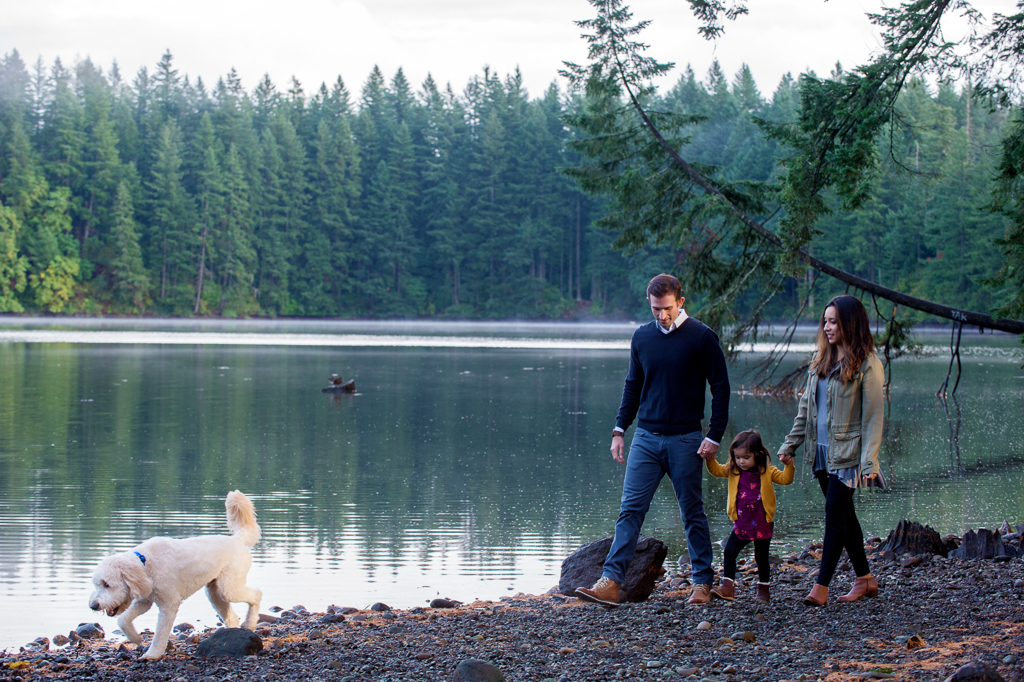 mom and dad hold a little girls hand as they walk along lacamas lake with their white labradoodle leading the way
