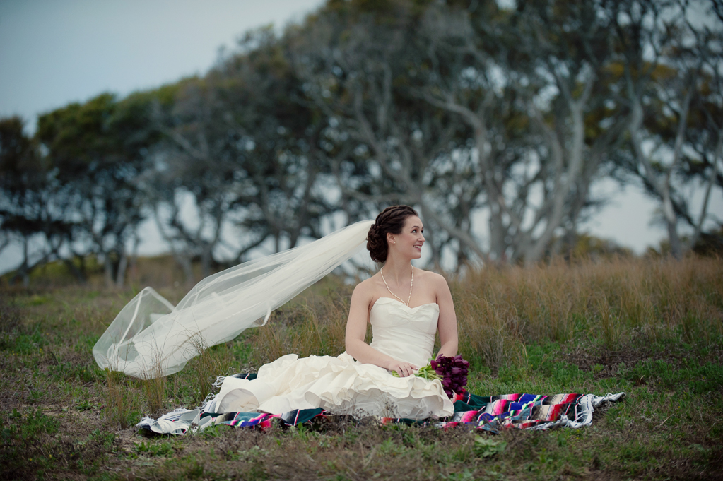 bride in wedding dress and long veil sits on a colorful mexican blanket in the grass in front of wind swept oak trees at fort fisher