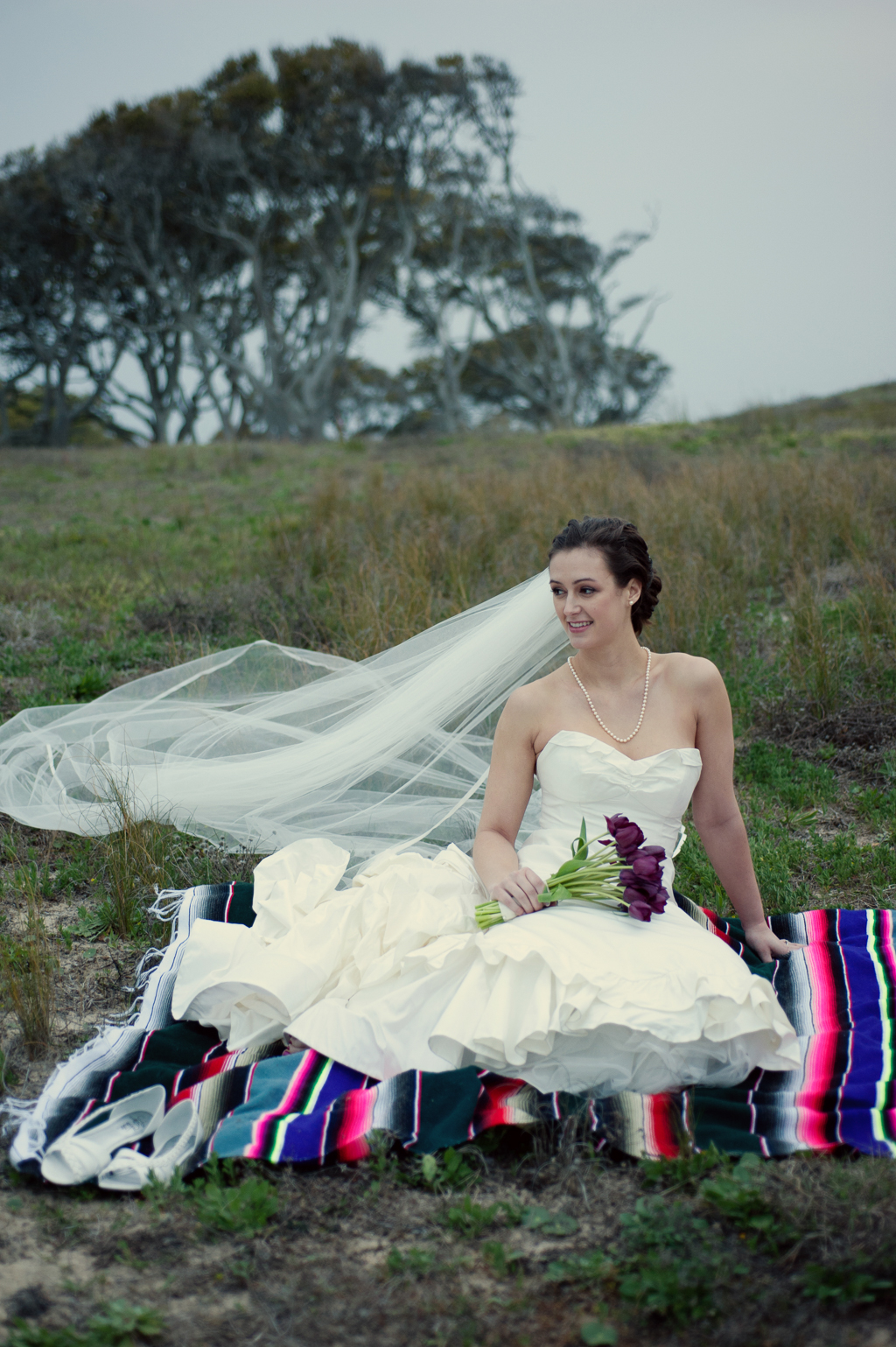 bride in wedding dress and long veil sits on a colorful mexican blanket in the grass in front of wind swept oak trees at fort fisher