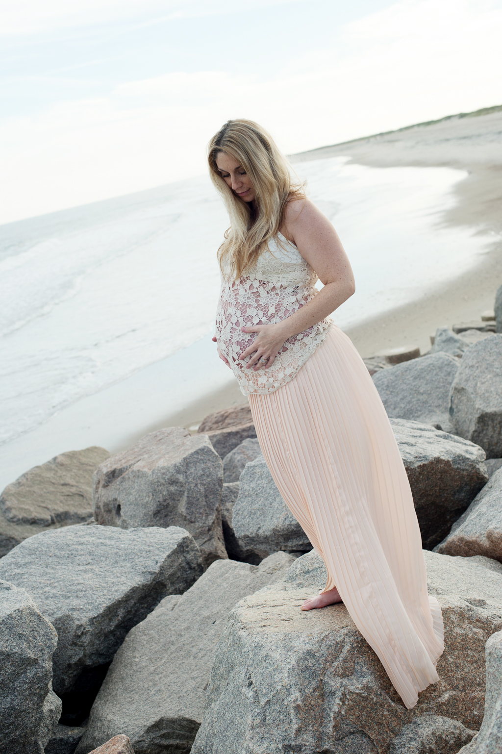 pregnant woman in long flowy skirt and lace top stands on the rocks at fort fisher