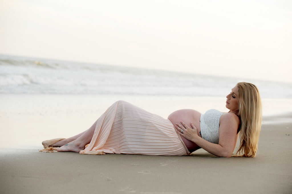 a pregnant woman in a long flowy skirt lays in the sand at the edge of the ocean as she looks out at the water while holding her belly