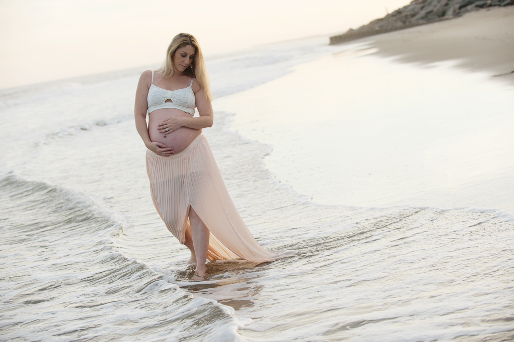 a pregnant woman in a long flowy skirt walks through the ocean's edge at fort fisher holding her belly