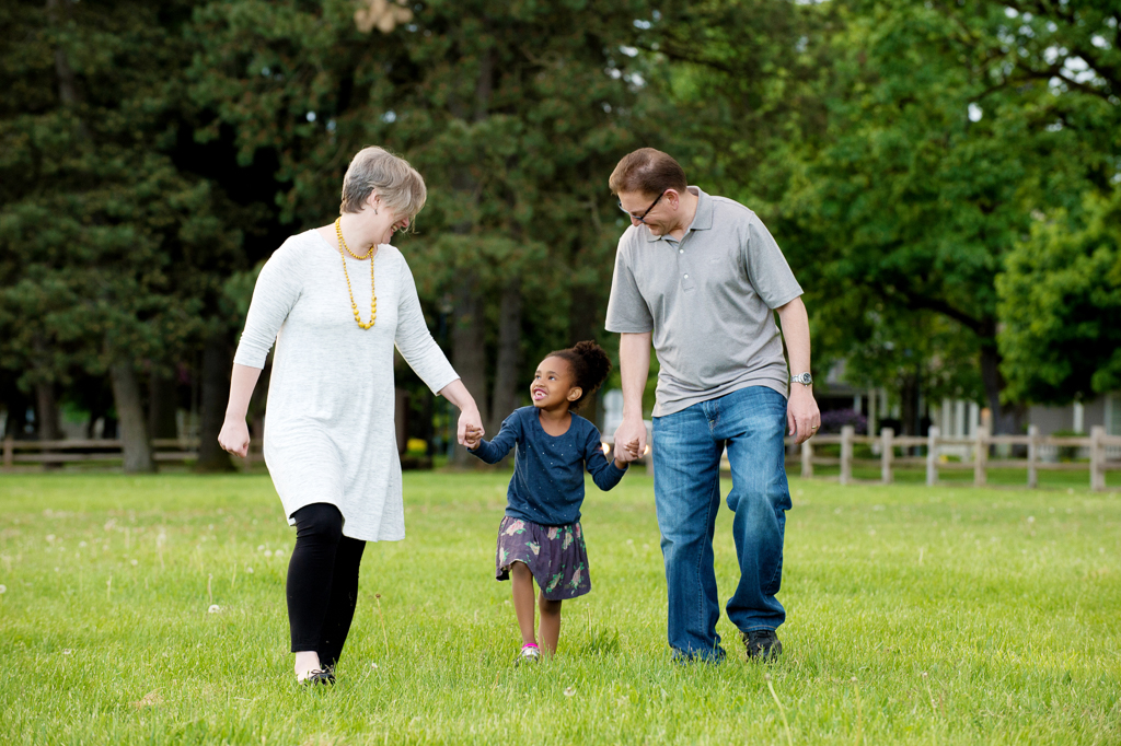 mom and dad hold hands with a young daughter as they walk through the yard at fort vancouver