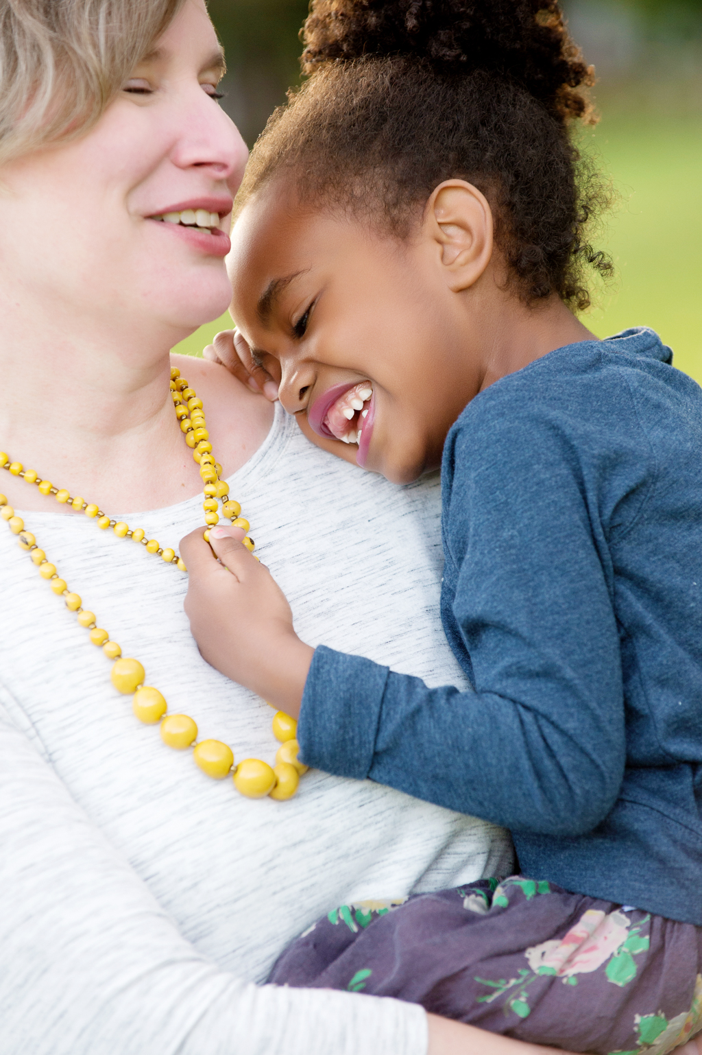 a young girl being held by her mom leans into her shoulder and laughs while holding her yellow necklace