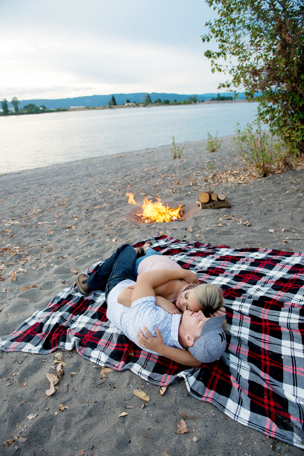 a man and woman cuddle on a plaid blanket on the beach next to a river with a campfire