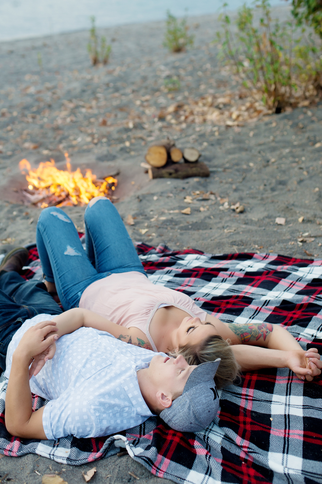 a man and woman cuddle on a plaid blanket on the beach next to a river with a campfire