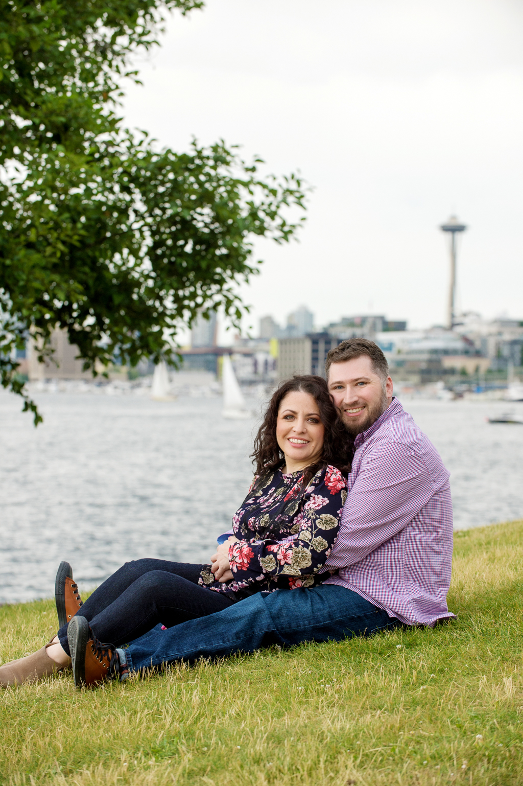 a couple sit in the grass in front of the waterfront with seattle space needle in the distance