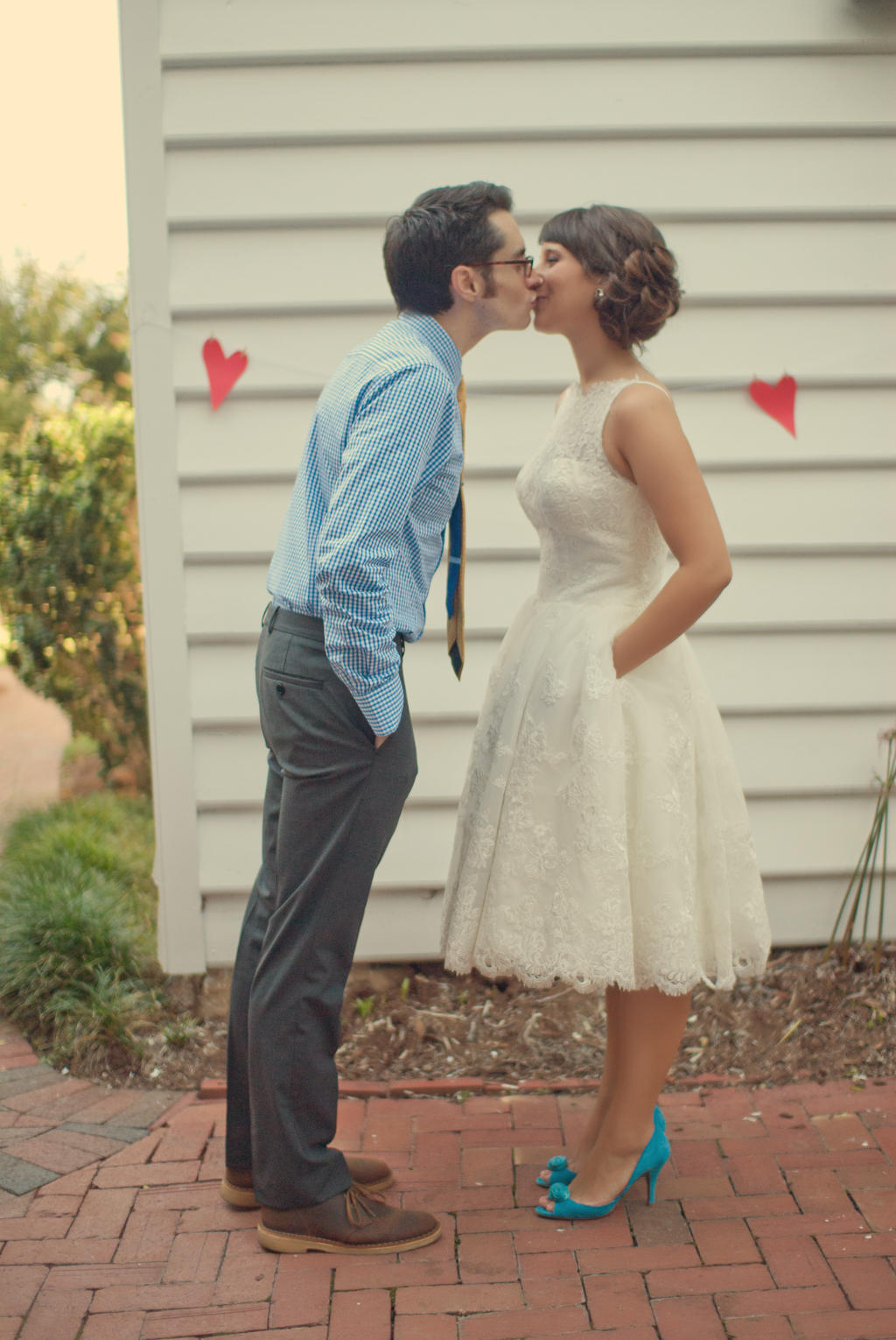 bride in tea length dress and blue shoes kisses groom in front of red hearts