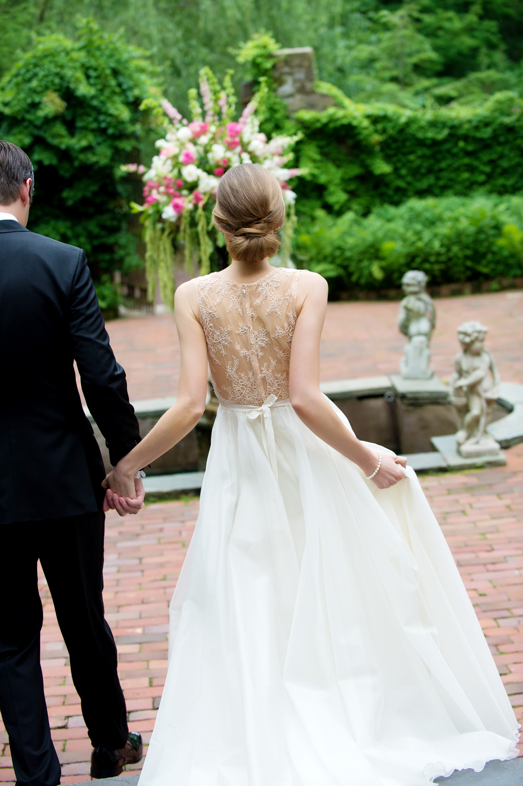 bride wears beautiful lace back wedding dress with pretty hair in bun as she walks towards fountain holding groom's hand
