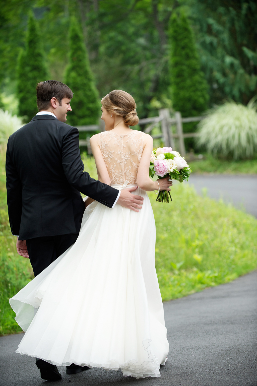 groom puts arm around bride as they walk down a path bride has pretty lace back wedding dress