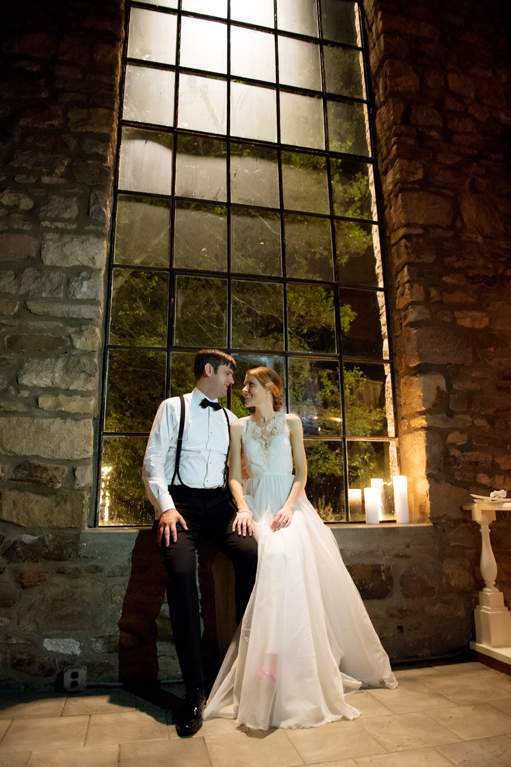 bride and groom sit in giant windows of holly hedge estate at night with candles lit