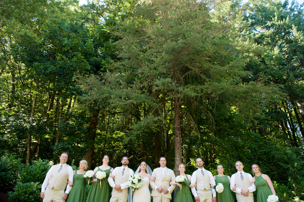 groomsmen in tan suits and bridesmaids in green dresses stand under tall trees with arms linked