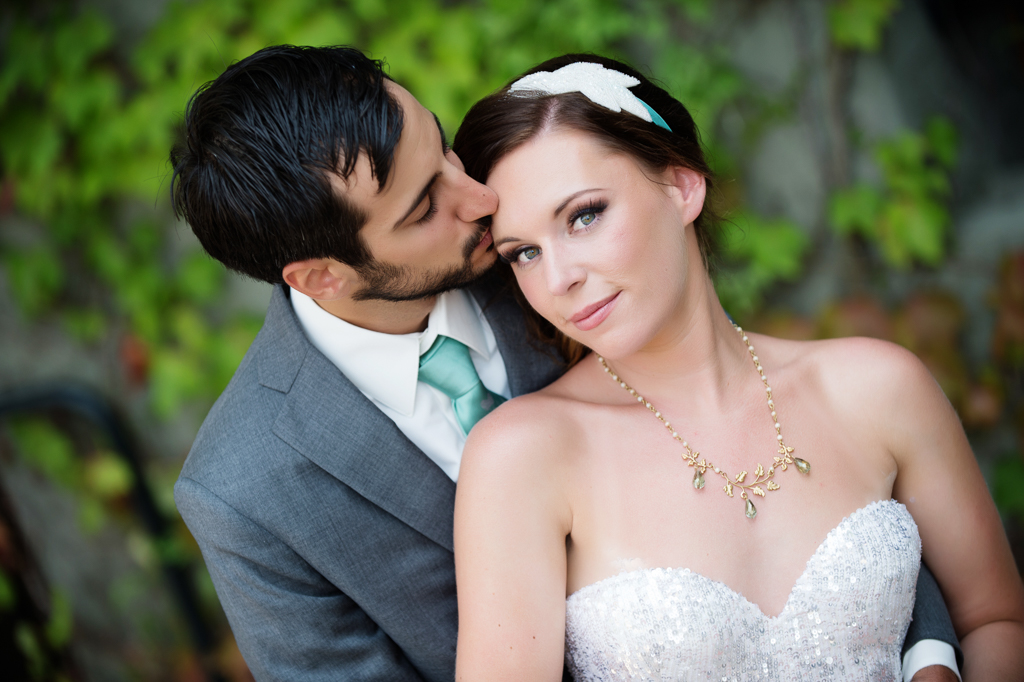 a bride with bright blue eyes looks intently at the camera as the groom kisses her forehead
