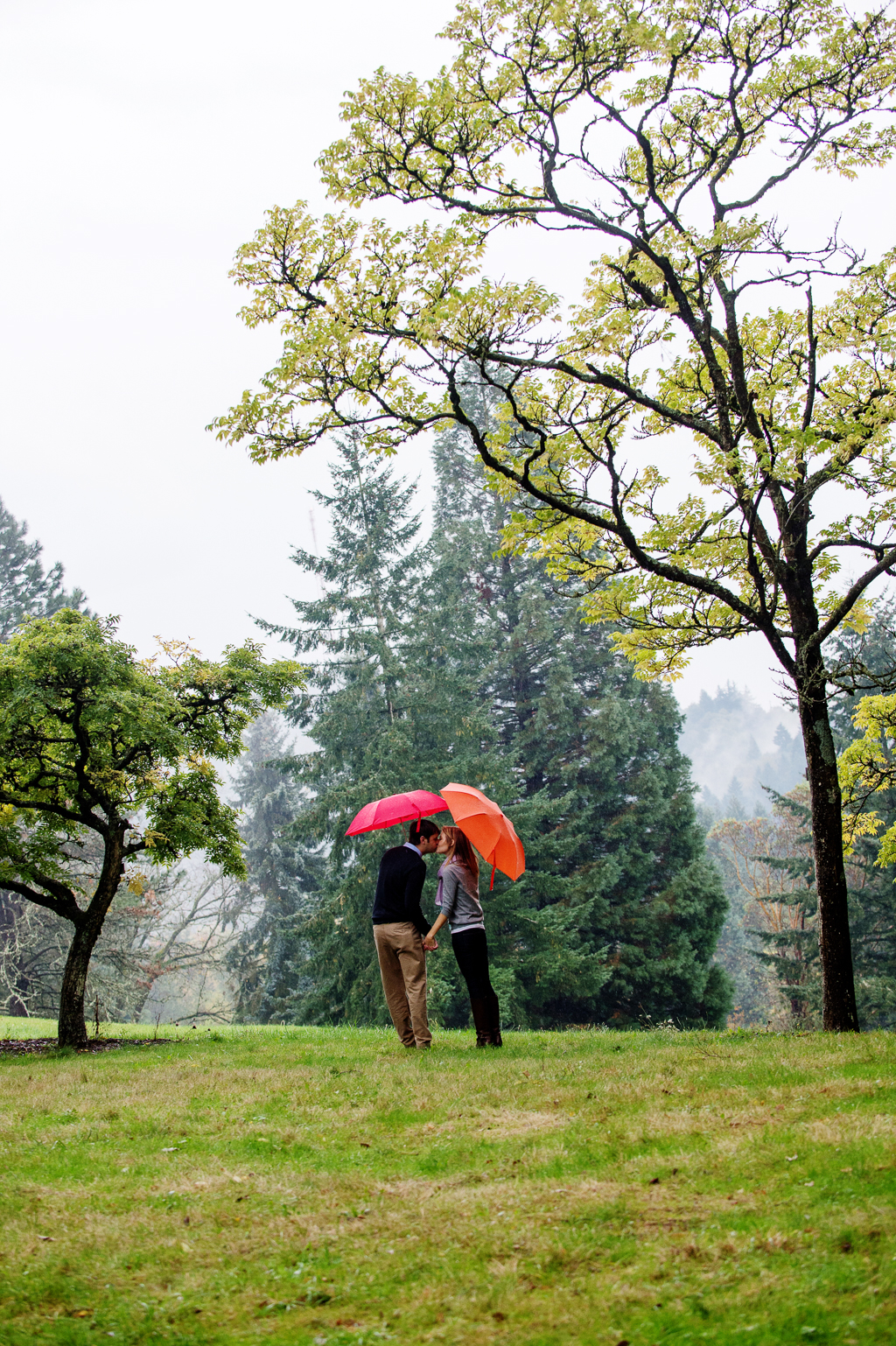 a girl with an orange umbrella kisses a man with a red umbrella under the trees at hoyt arboretum