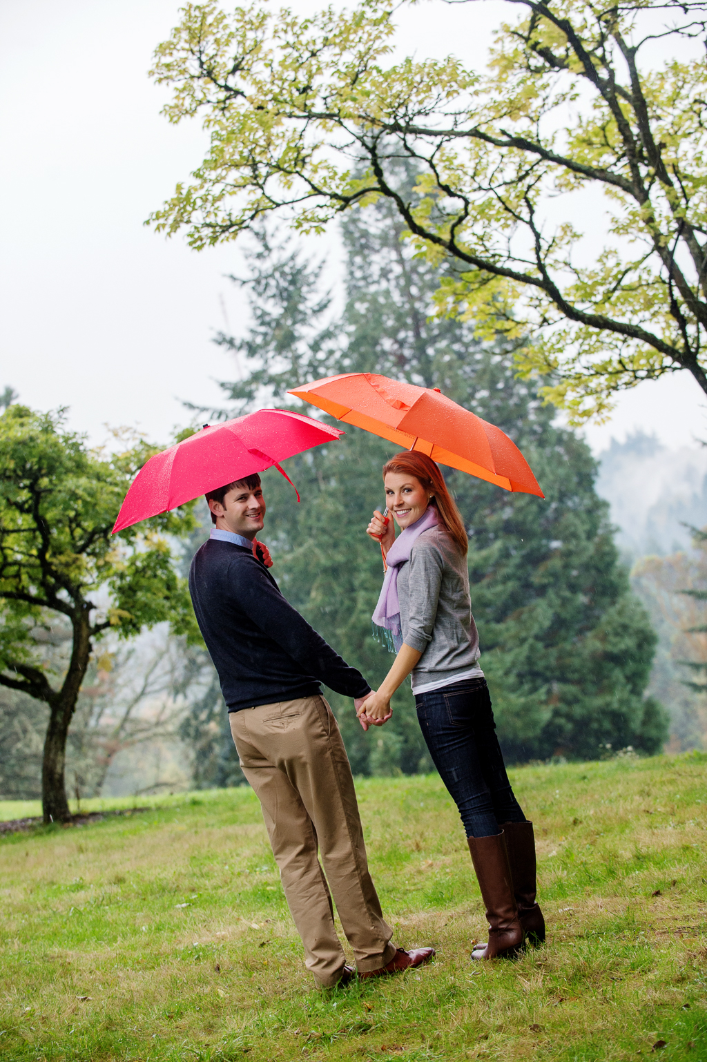 a girl with an orange umbrella walks with a man with a red umbrella under the trees at hoyt arboretum