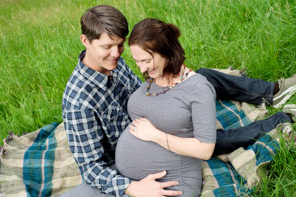 a man and a pregnant woman sit on a quilt in the grass and hug around the belly