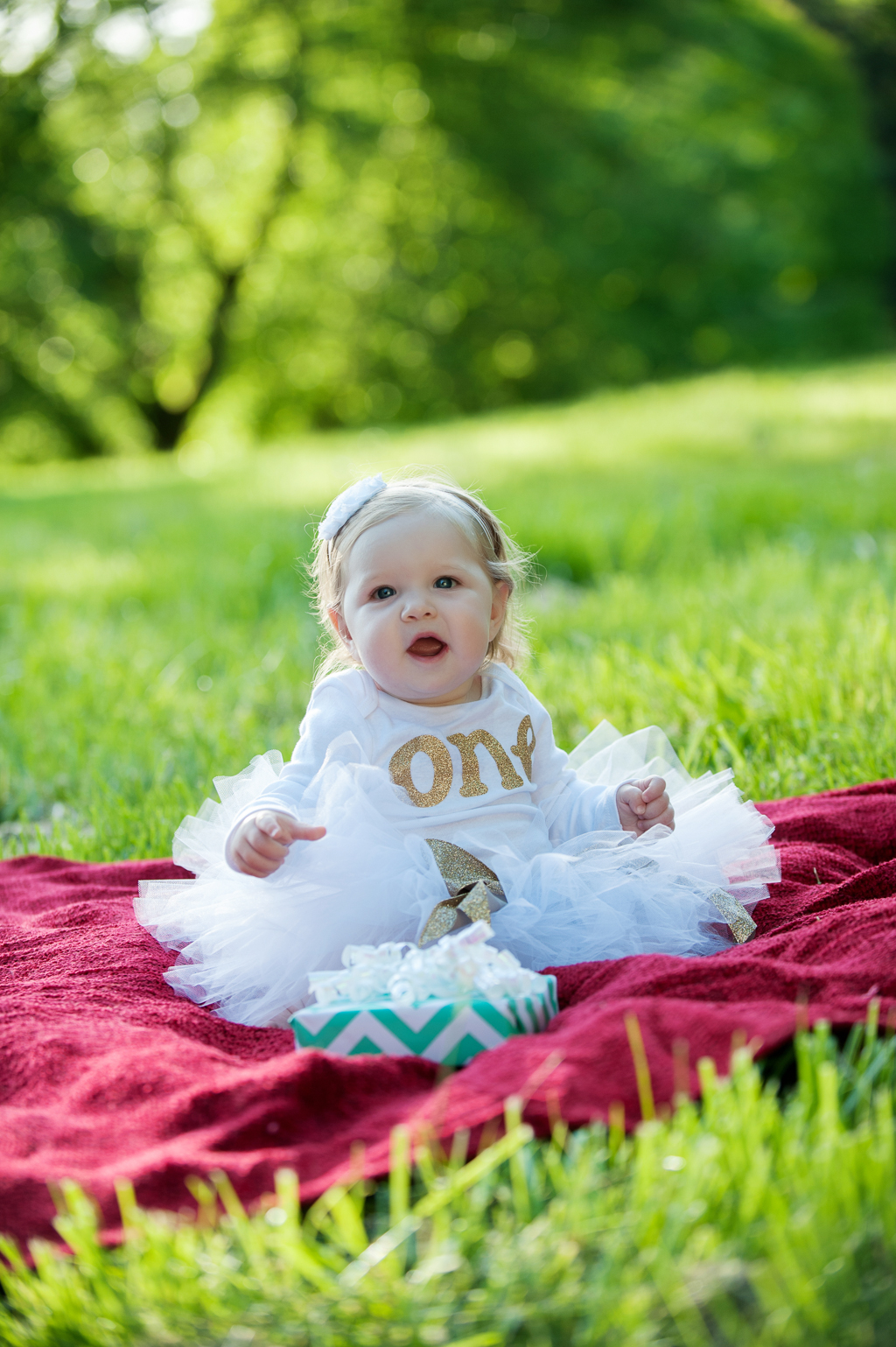a one year old baby girl sits on a blanket wearing a white tutu and holding a present