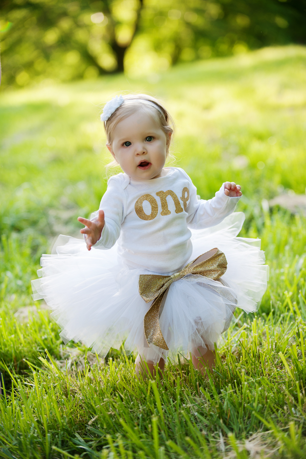 a baby girl wearing a white tutu with a big gold bow and a onesie that says one in gold letters stands in the grass