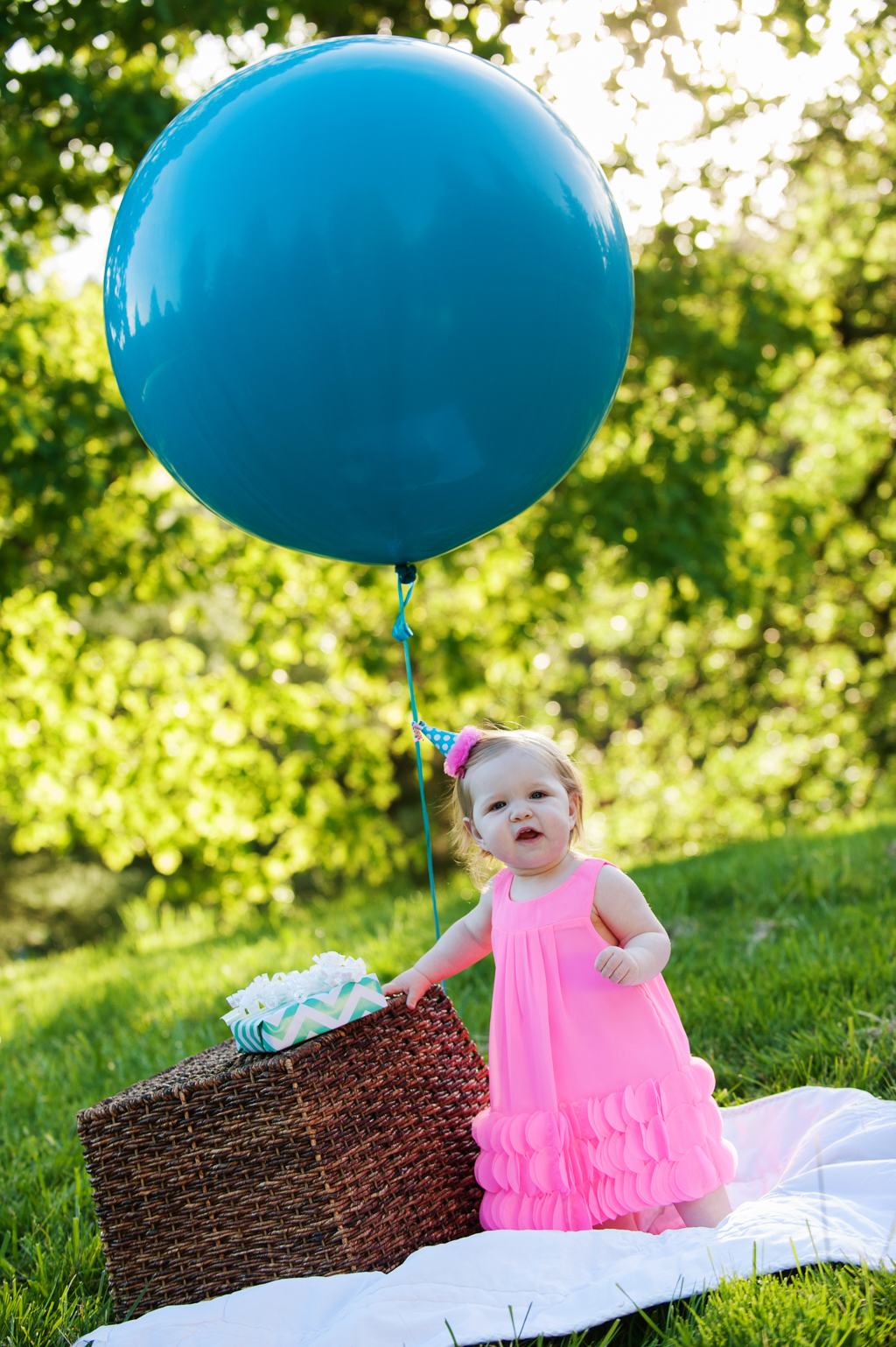 a baby girl wearing a hot pink dress stands in front of a birthday present and a giant blue balloon
