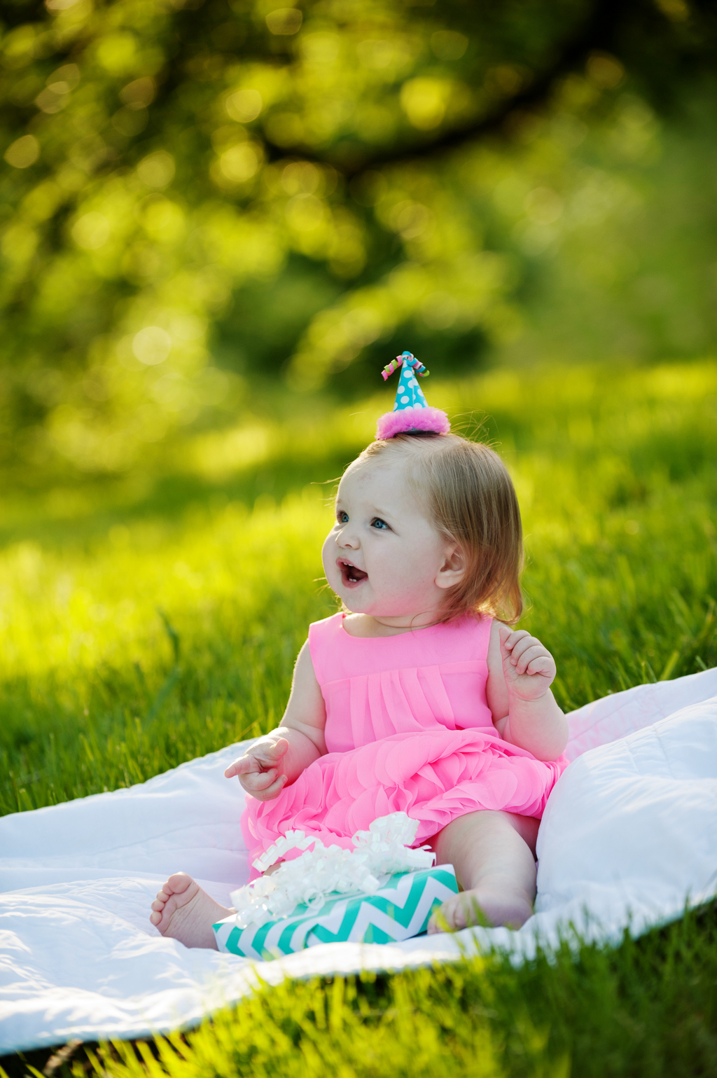 a baby girl wearing a hot pink dress and a tiny birthday hat sits in the grass holding a present