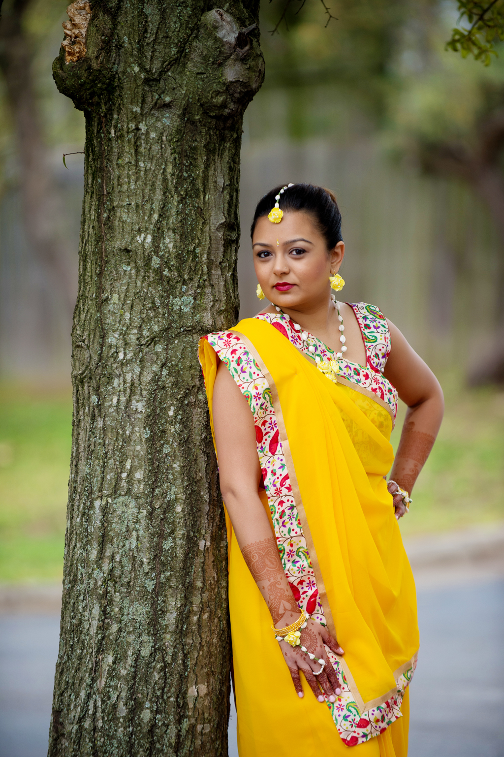 an indian bride dressed in a bright yellow sari leans against a tree
