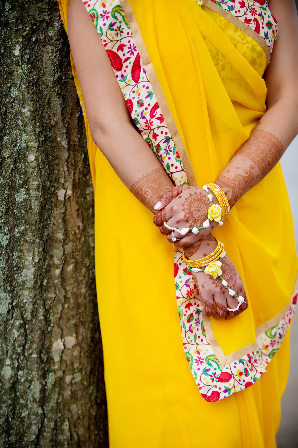 an indian bride dressed in a bright yellow sari leans against a tree