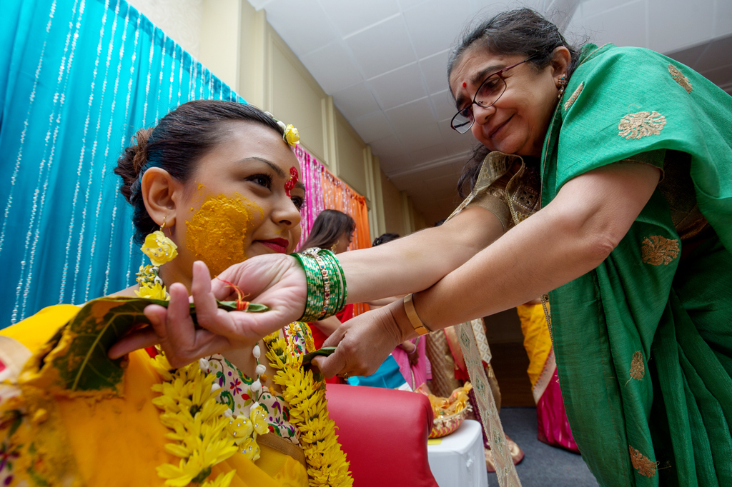an indian bride dressed in a bright yellow sari has turmeric paste applied to her face during ceremony