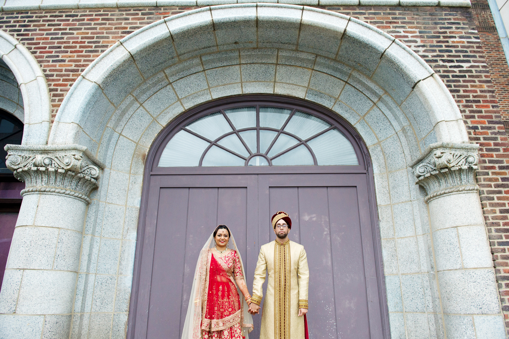 bride and groom in traditional red and gold indian wedding attire hold hands in front of large purple doors