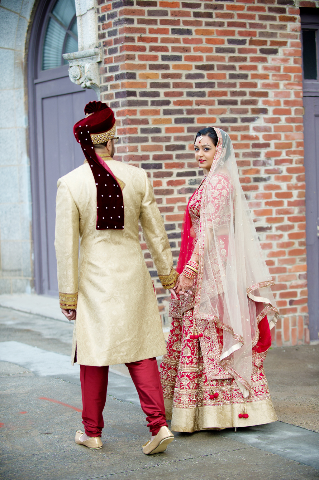 bride and groom in traditional red and gold indian wedding attire hold hands