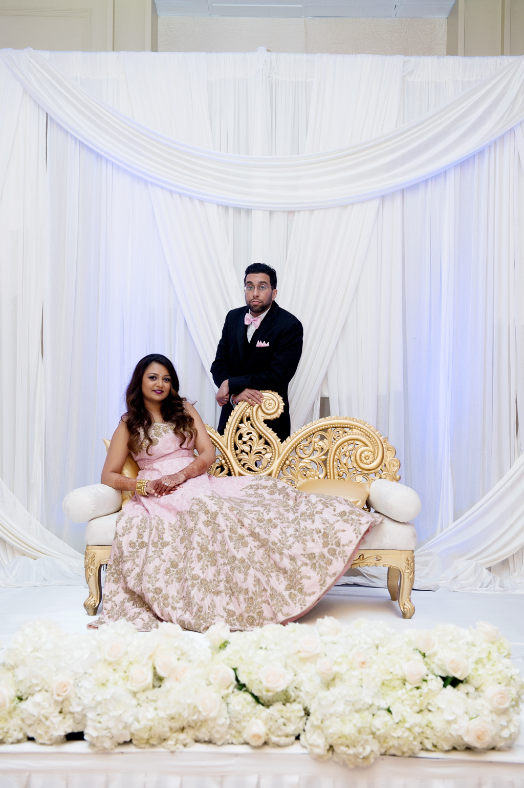 an indian bride wears a pink and gold dress and lounges on an intricate gold couch as her groom stands over her