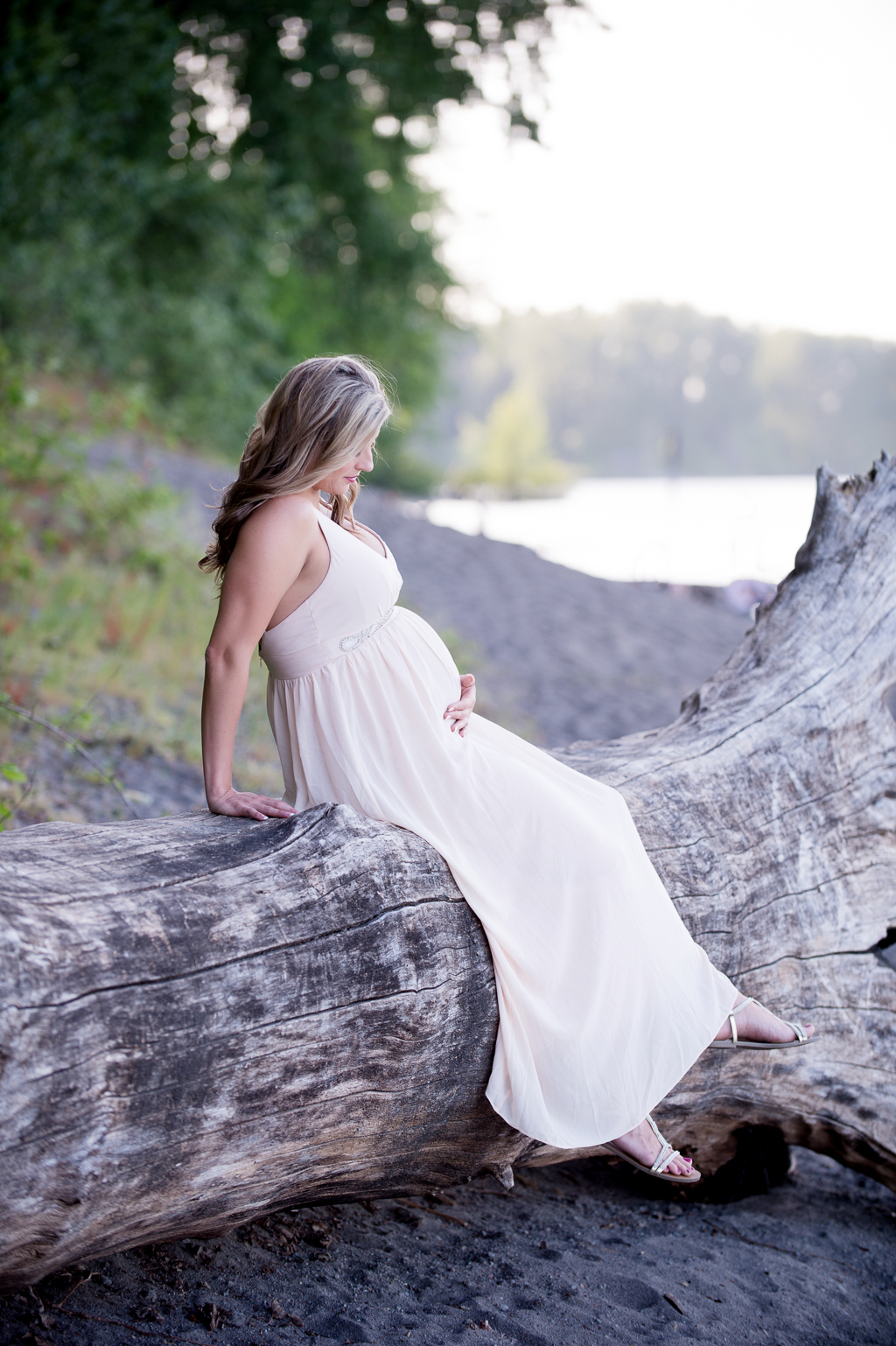 pregnant woman in pink dress sits on a fallen log by the river and looks at her belly